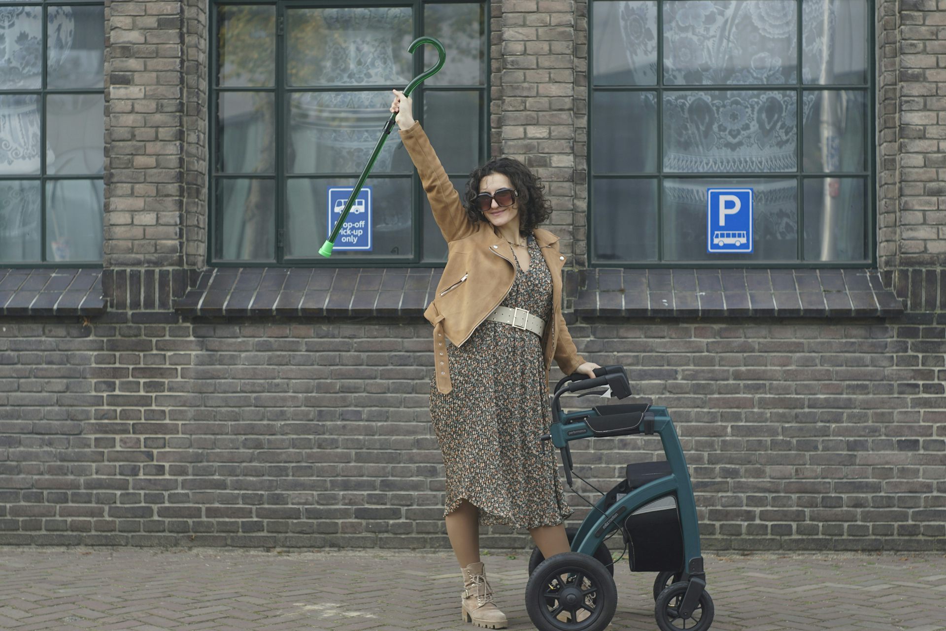 A young woman on a sidewalk, holding up a cane and leaning on a wheeled walker.