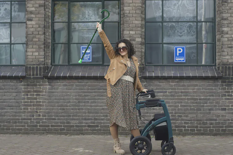 A young woman on a sidewalk, holding up a cane and leaning on a wheeled walker.