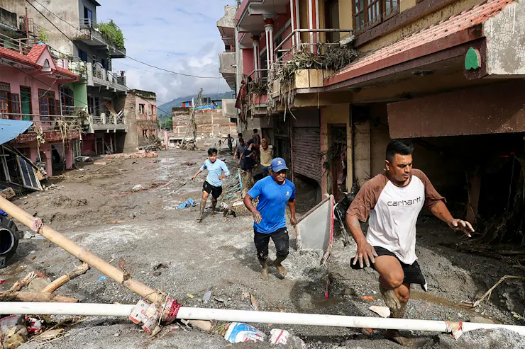 Three men running through a muddy street barefoot. Muddy vegetation is stuck to the second-floor balconies and mud can be seen most of the way up the side of a three-story building.