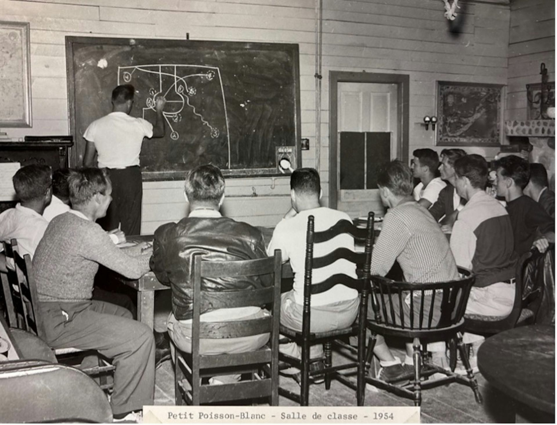 Photo en noir et blanc d’un enseignant écrivant à la craie au tableau devant un groupe de jeunes hommes