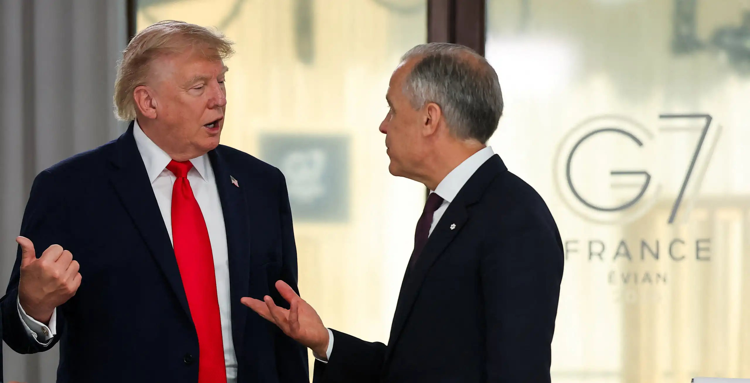 A large man with fluffy blonde-ish hair gestures while speaking to a slender man with short grey hair whose left hand is raised while speaking.