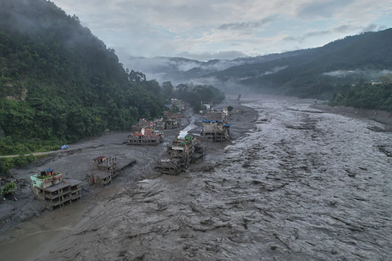 A torrent of muddy floodwater rushes through a Nepalese valley, with houses along the banks and lush green hills and mist-covered mountains in the background.