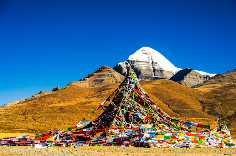 A snow-capped mountain rises among a range of peaks, with Buddhist prayer flags lining the path leading toward it.
