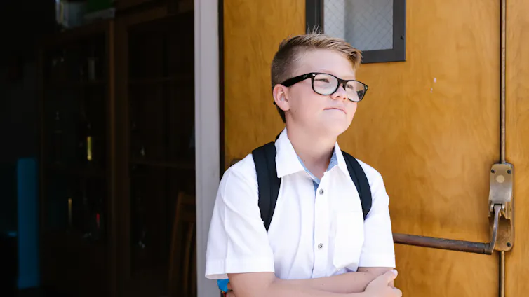 A school student in a white shirt and backpack stands with their arms folded.
