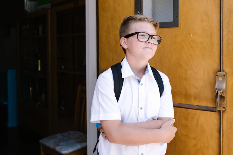 A school student in a white shirt and backpack stands with their arms folded.