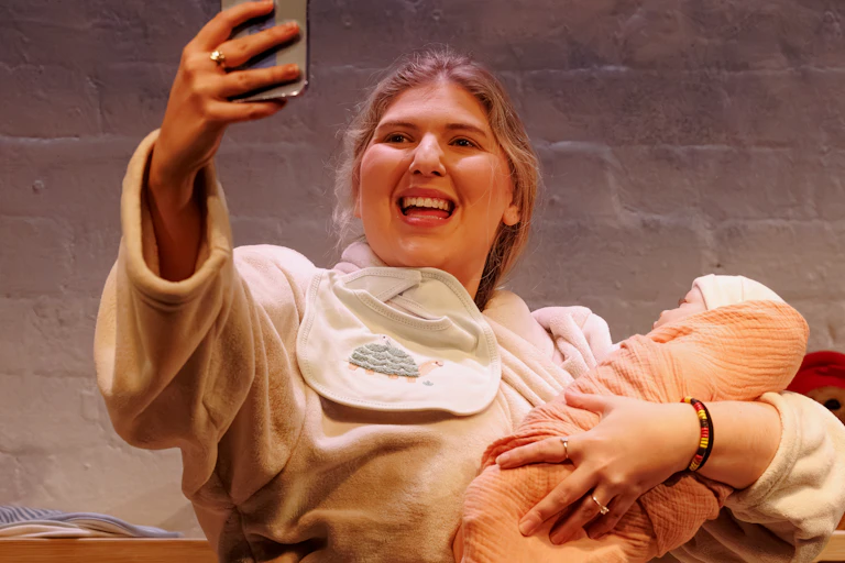A young woman with blonde hair wearing a bathrobe and holding a lifelike baby doll takes a selfie while smiling.