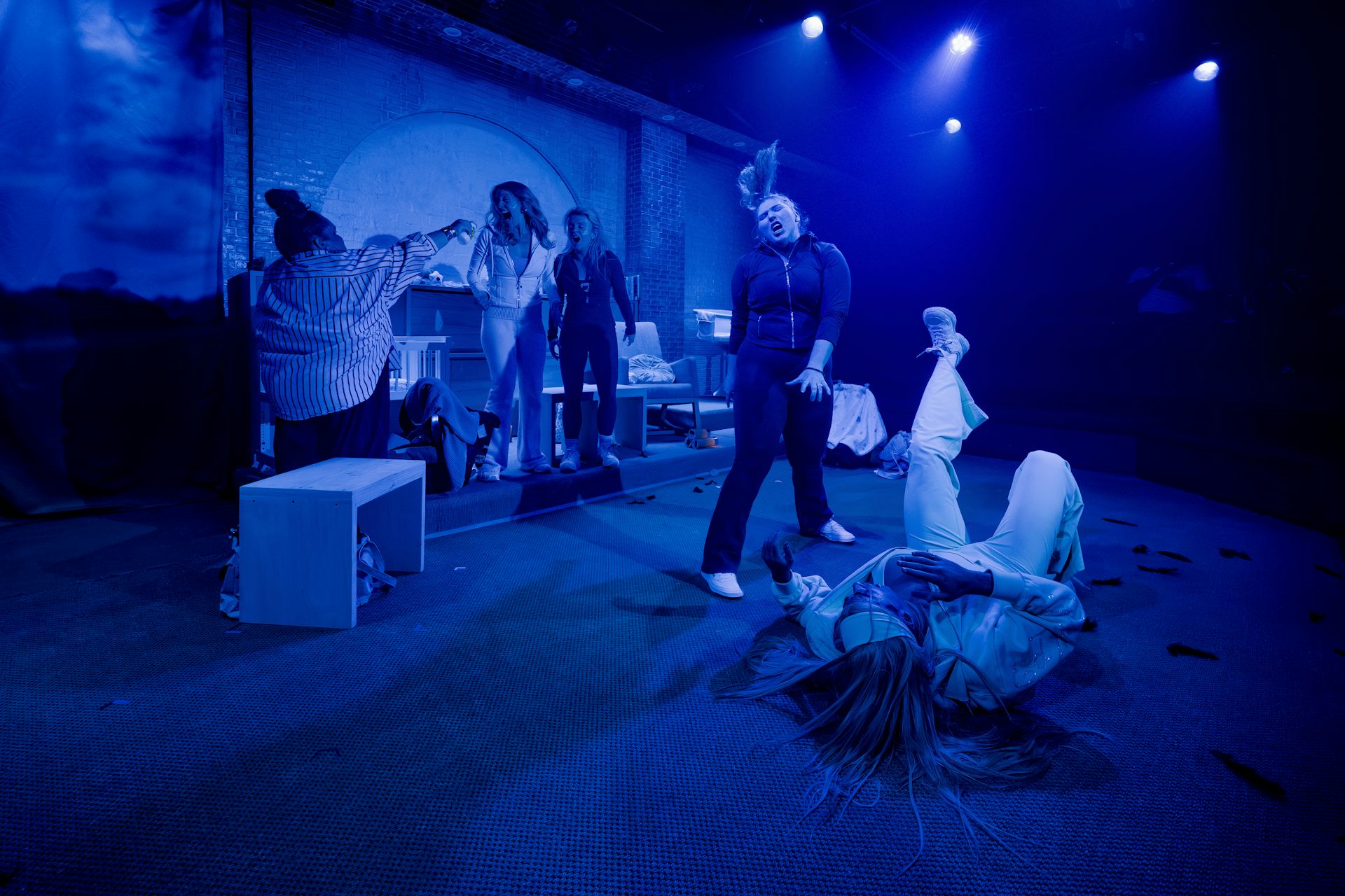 Five performers in various positions on a blue-lit stage, including one lying down on the floor and one dancing.