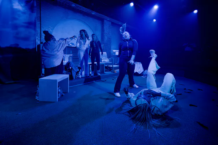 Five performers in various positions on a blue-lit stage, including one lying down on the floor and one dancing.