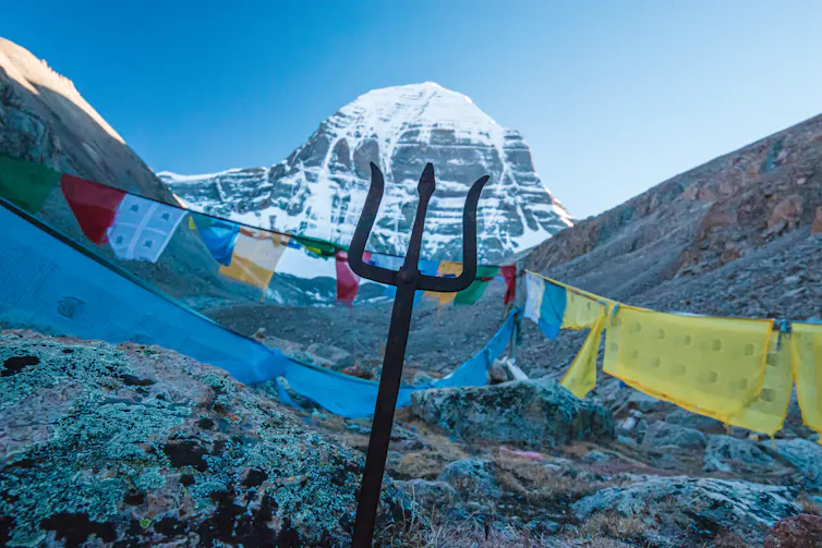 Colourful prayer flags and a trident, symbolizing the Hindu god Shiva, placed in front of a snow-capped mountain.