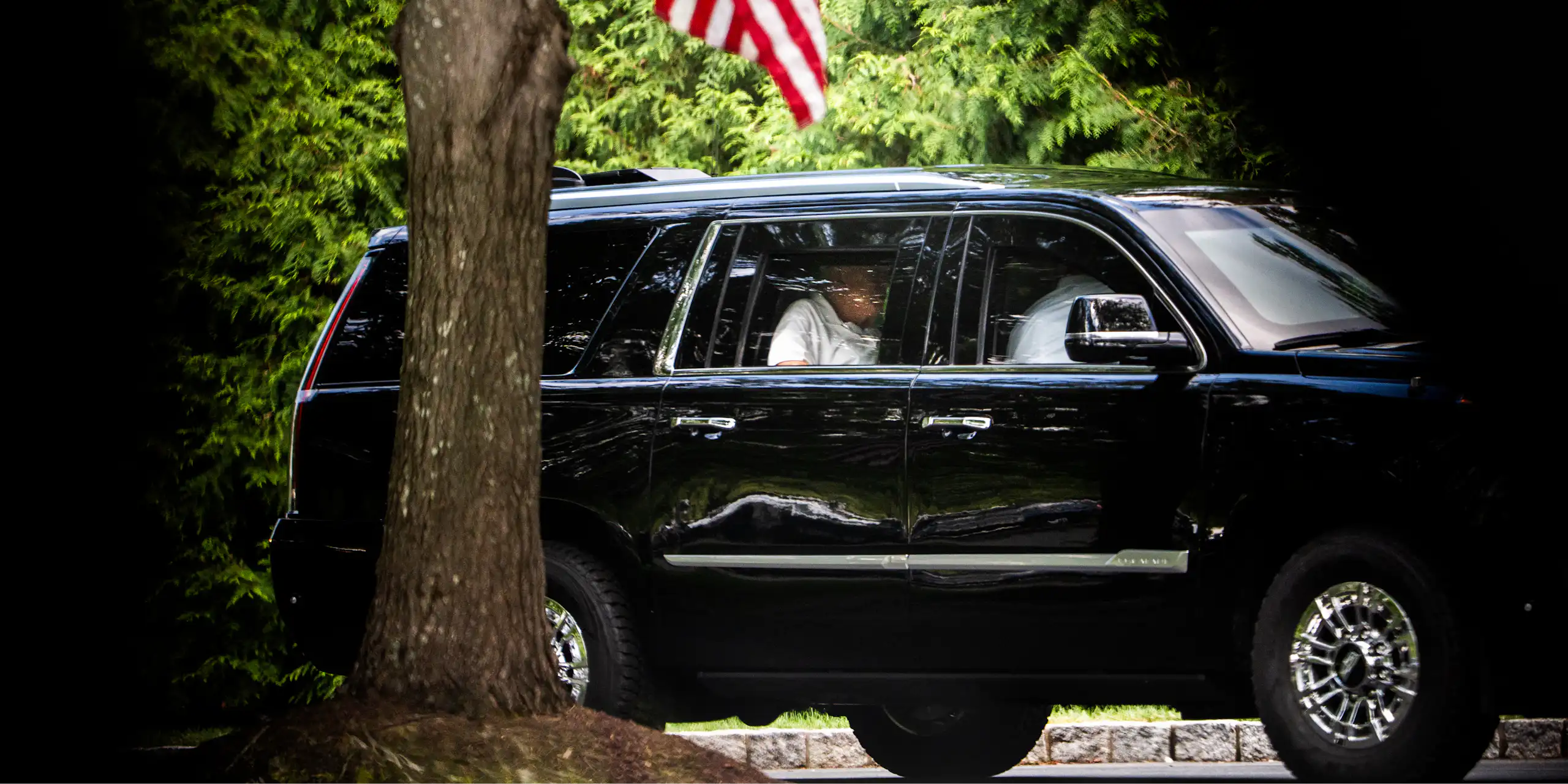 A rotund man in a white polo shirt in the back of a black SUV. An American flag hangs from a tree overhead.