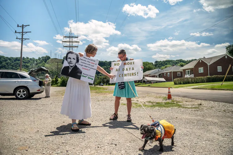Two people stand in gravel parking area and hold signs protesting data centers. One signs says 'No more AI data centers!'