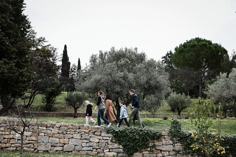 Une famille de trois enfants avec leurs deux parents marche dans un parc planté d’oliviers.