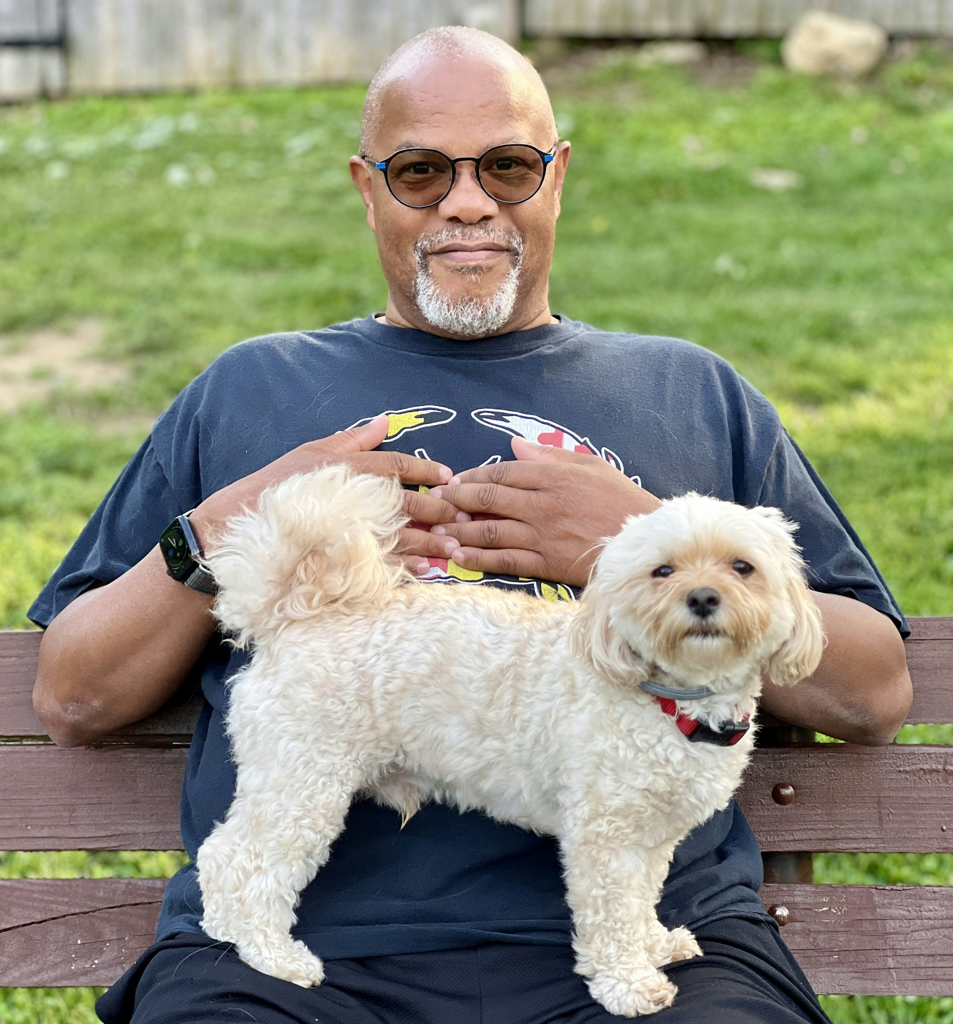 A man in sunglasses sits on park bench with small white dog standing on his lap