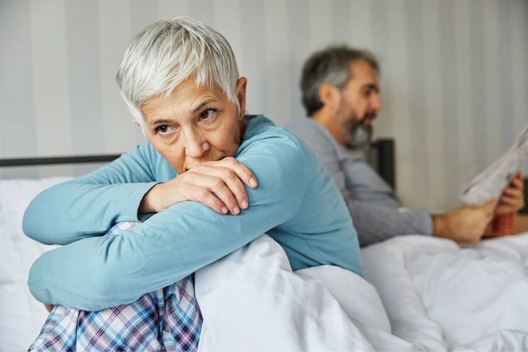 middle-aged woman sitting on the edge of her bed looking sad while her husband reads the paper behind her.