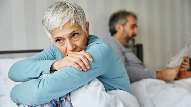 middle-aged woman sitting on the edge of her bed looking sad while her husband reads the paper behind her.
