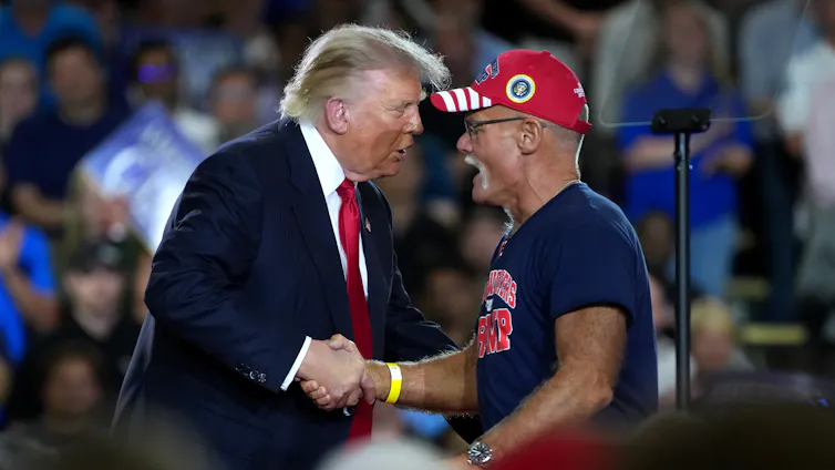 A man in suit and tie shakes hands with a man wearing a ball cap on stage.