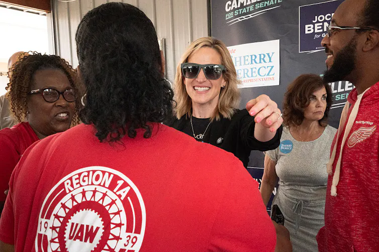 A woman wearing sunglasses smiles at a woman wearing a red t-shirt.
