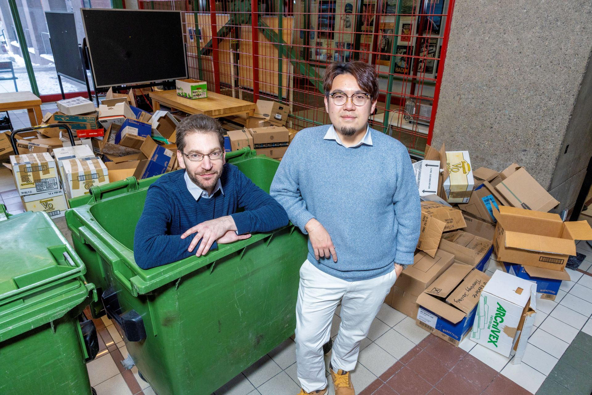 Two men pose with green waste bins and piles of cardboard