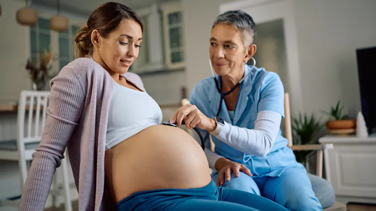 Visiting nurse examining pregnant woman's belly with a stethoscope at home