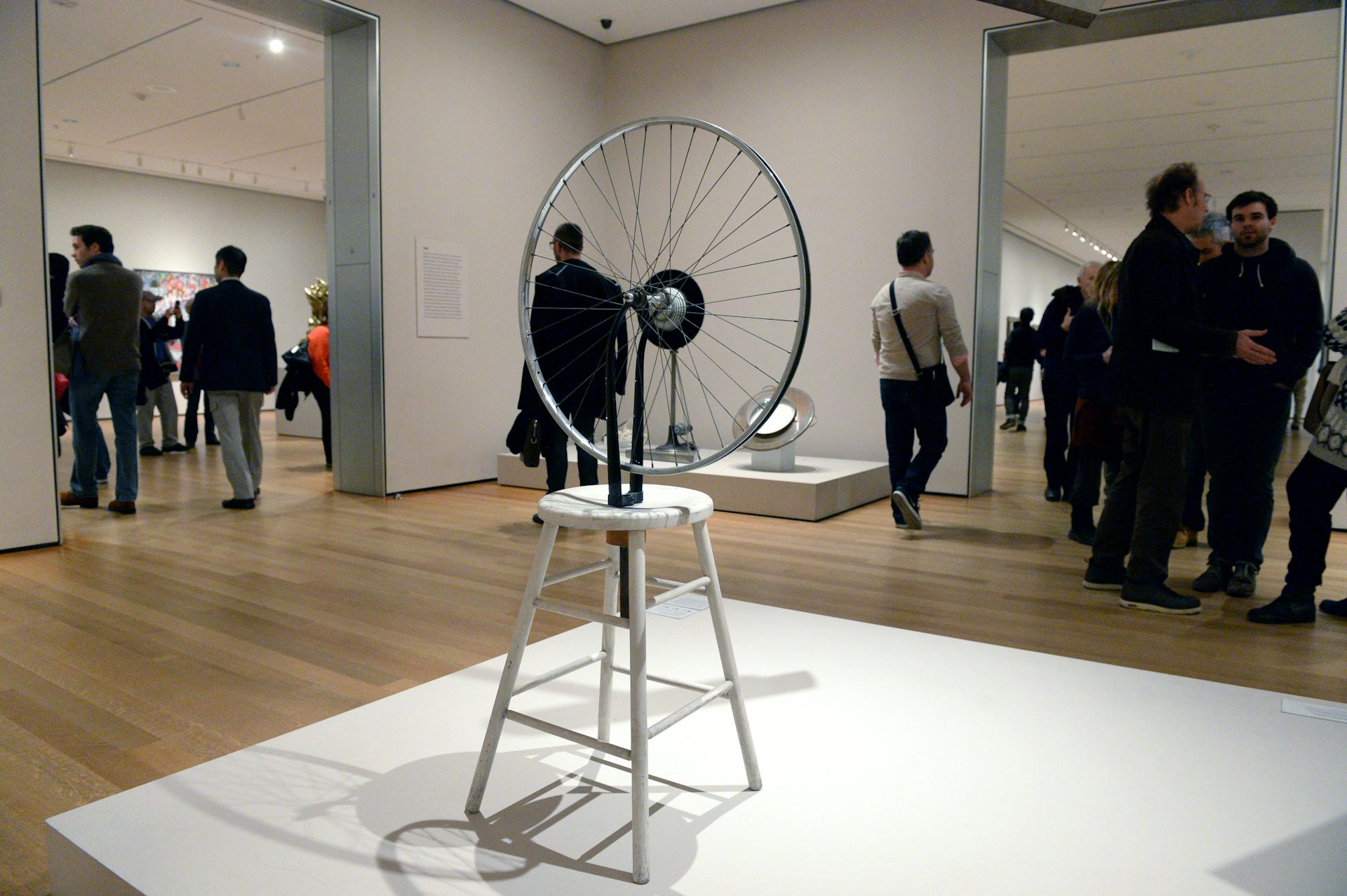 Sculpture of bicycle wheel on a white stool in a gallery setting with people in background