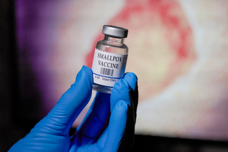 Blue gloved hand holding a vial of smallpox vaccine in front of the monkeypox virus graphic on a monitor