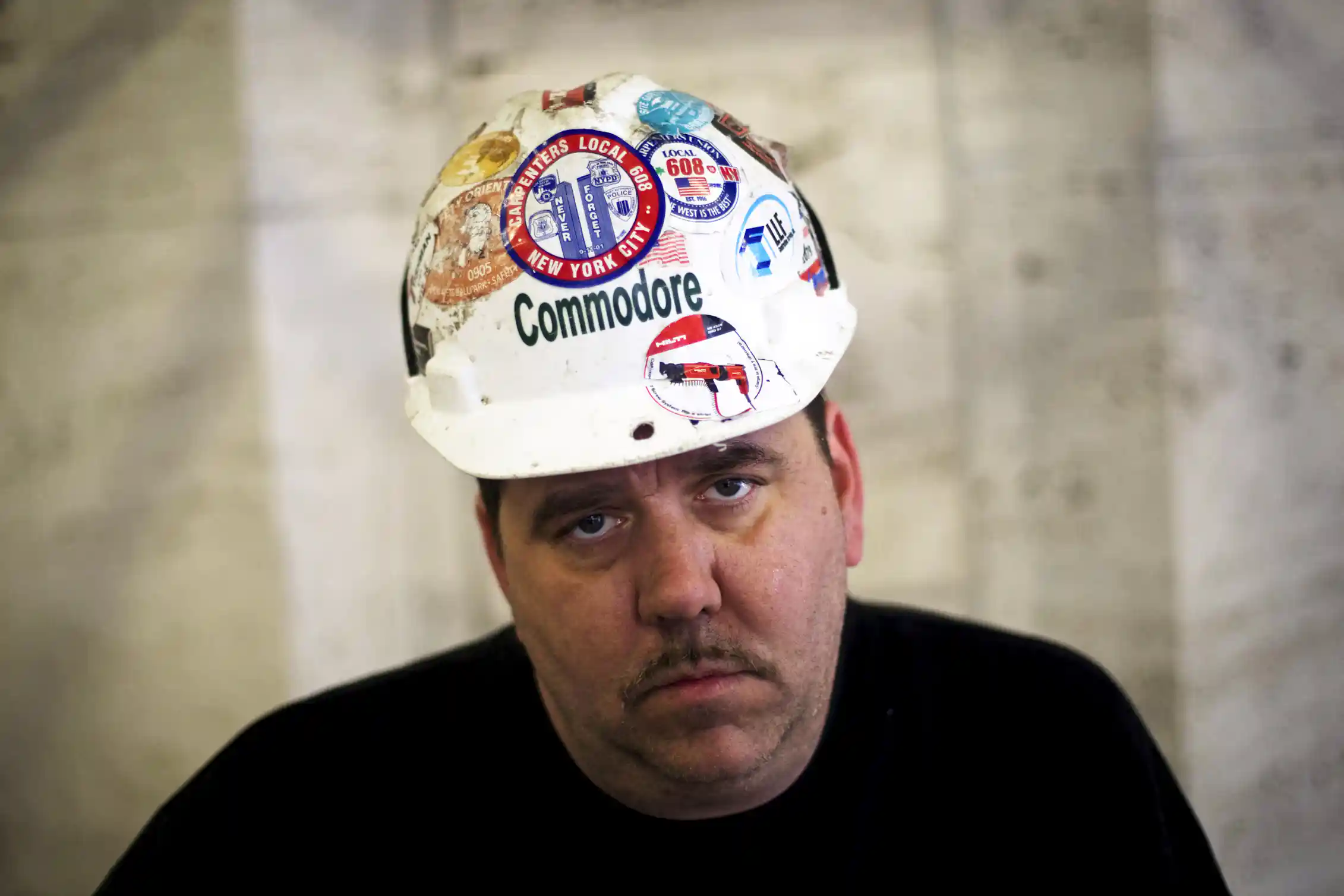 Persona que mira directamente a la cámara, con un casco de construcción adornado con varios parches, entre ellos uno con un grabado de las Torres Gemelas y la l