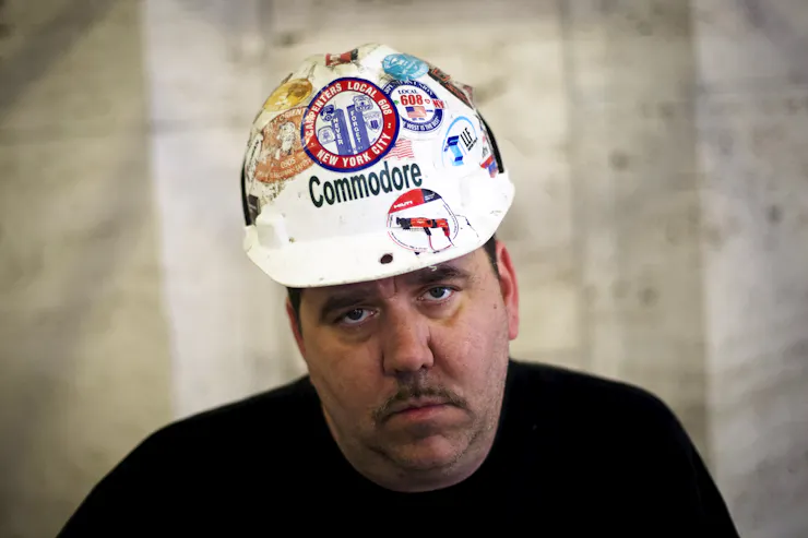 Person looking directly at the camera, wearing construction hat adorned with various patches, including one with an etching of the Twin Towers reading 'NEVER FORGET'