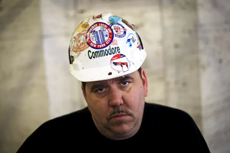 Person looking directly at the camera, wearing construction hat adorned with various patches, including one with an etching of the Twin Towers reading 'NEVER FORGET'