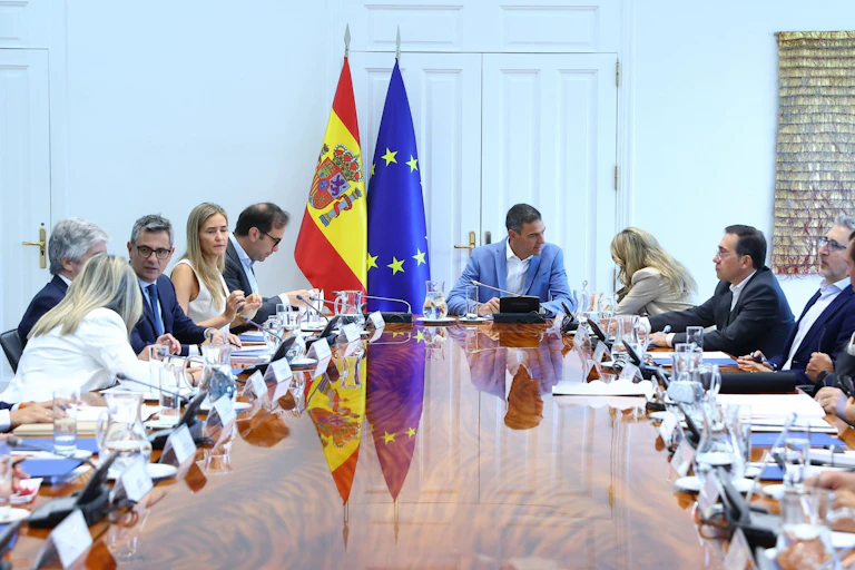 Varias personas sentadas en torno a una mesa con las banderas de España y la UE al fondo.