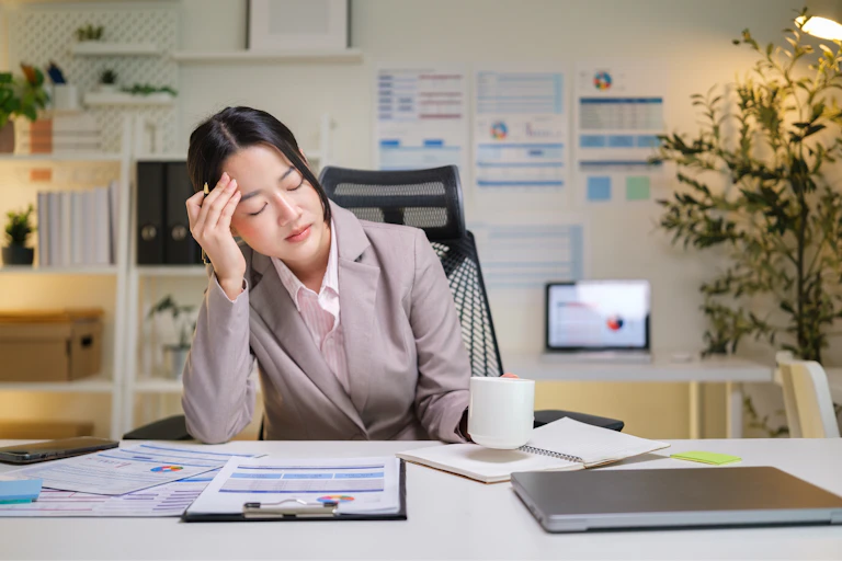 A tired business woman leans her head against her hand while resting her eyes at her desk.