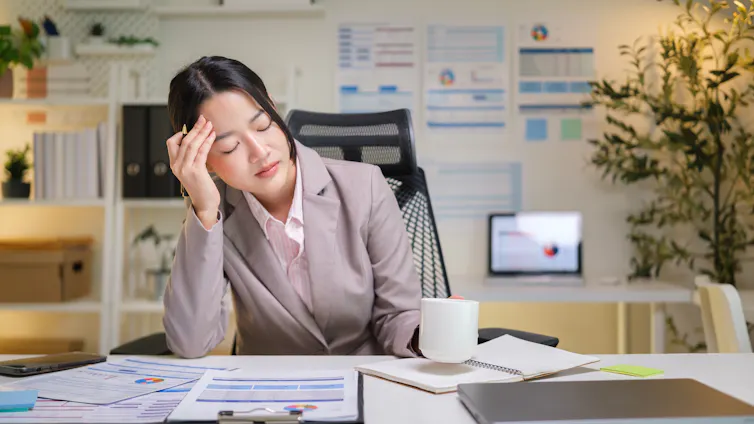 A tired business woman leans her head against her hand while resting her eyes at her desk.