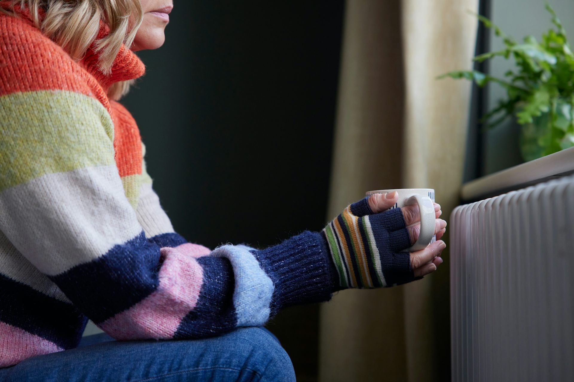 A woman wearing gloves and holding a hot drink next to a radiator