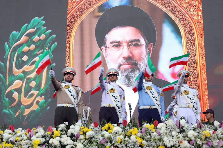 Four men in uniforms wearing sashes in Arabic writing stand in front of a banner of Iran's supreme leader, Mojtaba Khamenei.