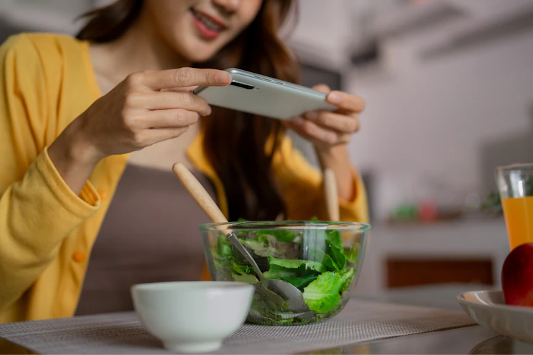 Une jeune femme prend en photo avec son smartphone le bol de salade qu'elle a devant elle.
