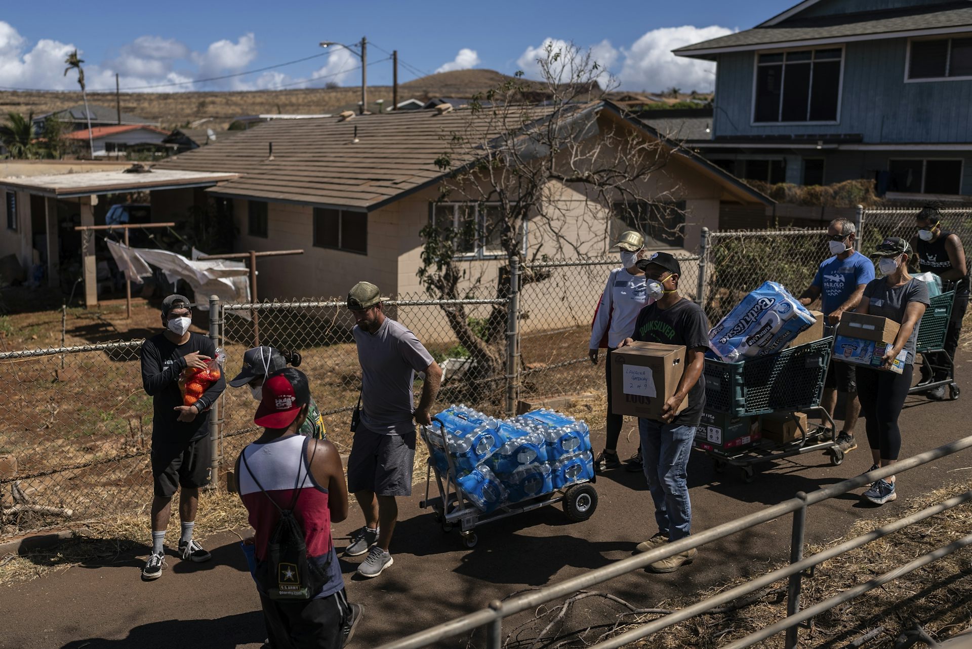 Eight people walk down a narrow road, carrying boxes of water and other supplies.