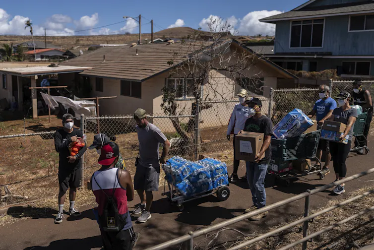 Eight people walk down a narrow road, carrying boxes of water and other supplies.