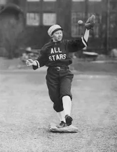 Black-and-white photo of a woman wearing a cap and a baggy uniform reading 'ALL STARS' while standing on a base and catching a ball.