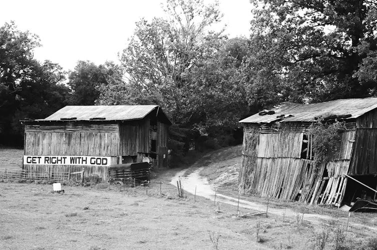 A black-and-white photograph of two dilapidated barns, one of which has 'Get right with God' painted on its side.
