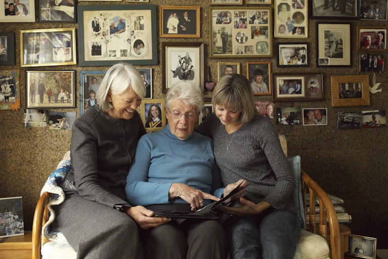 Three generations of women look at a photo album together while behind them a wall features dozens of framed photos.
