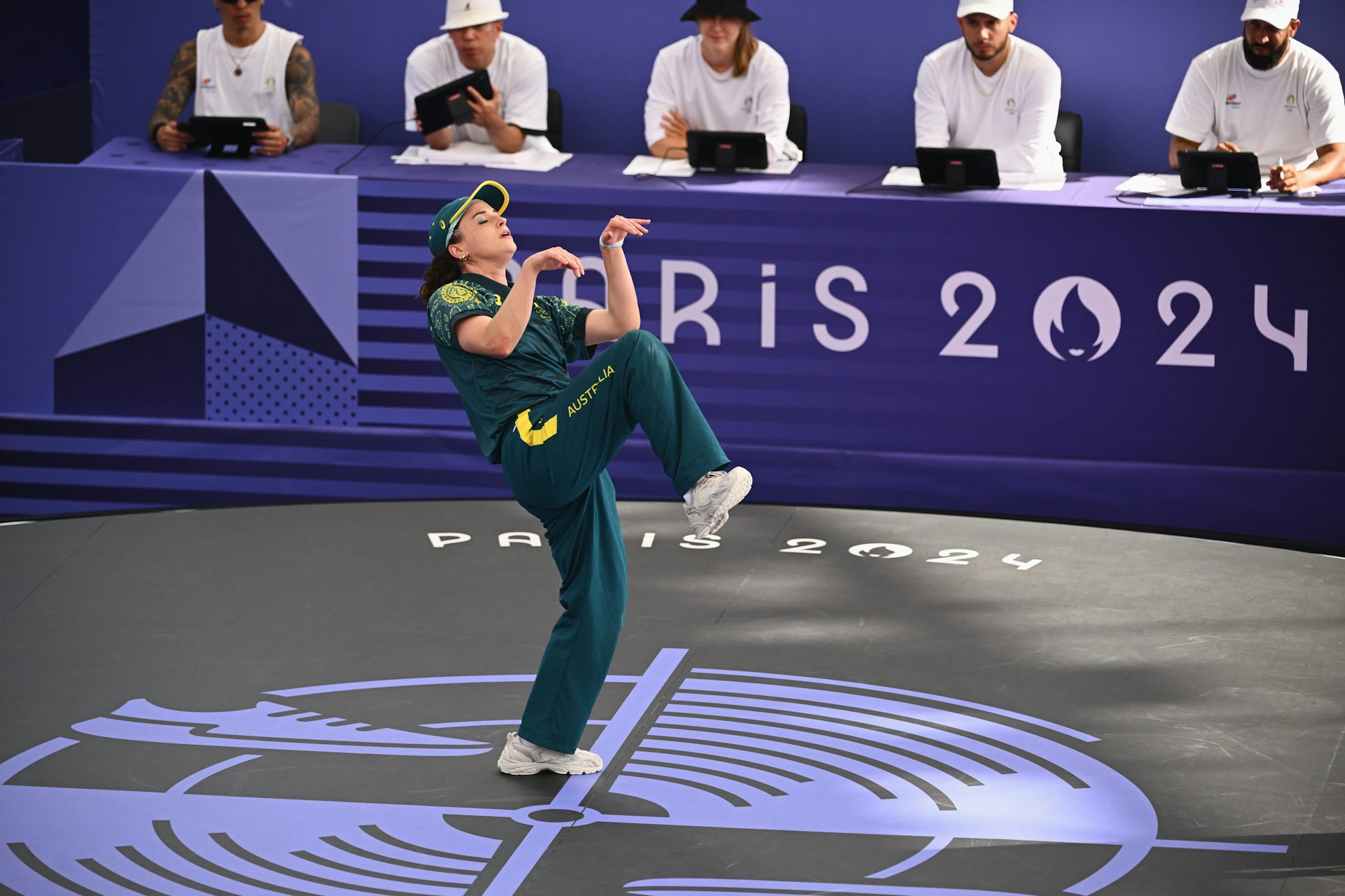 A woman in a green tracksuit does a kangaroo pose on an Olympic stage, with judges in the background.