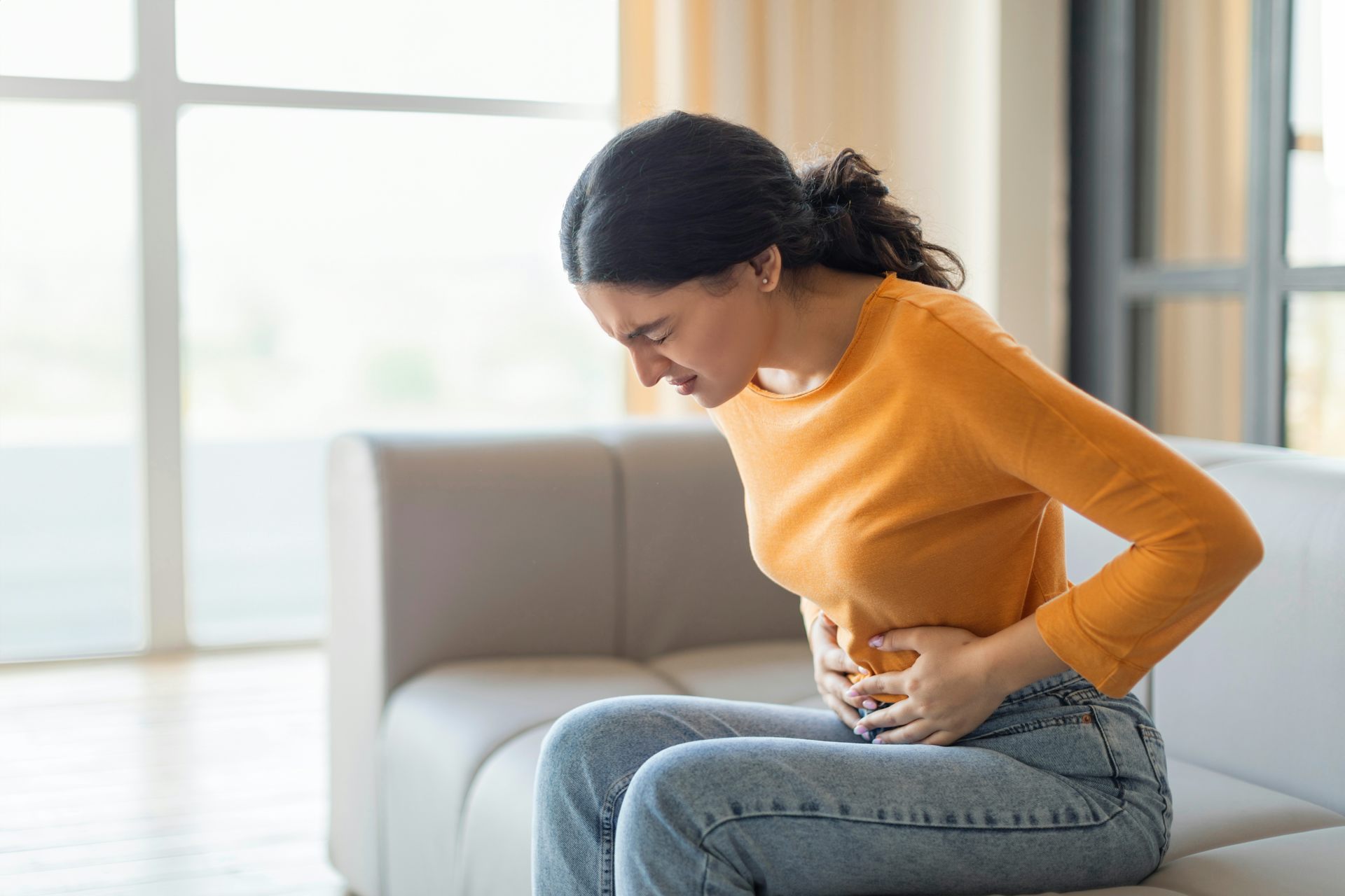 A young woman sitting on a couch holds her stomach in pain.