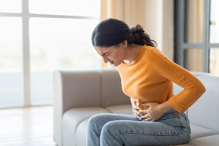 A young woman sitting on a couch holds her stomach in pain.
