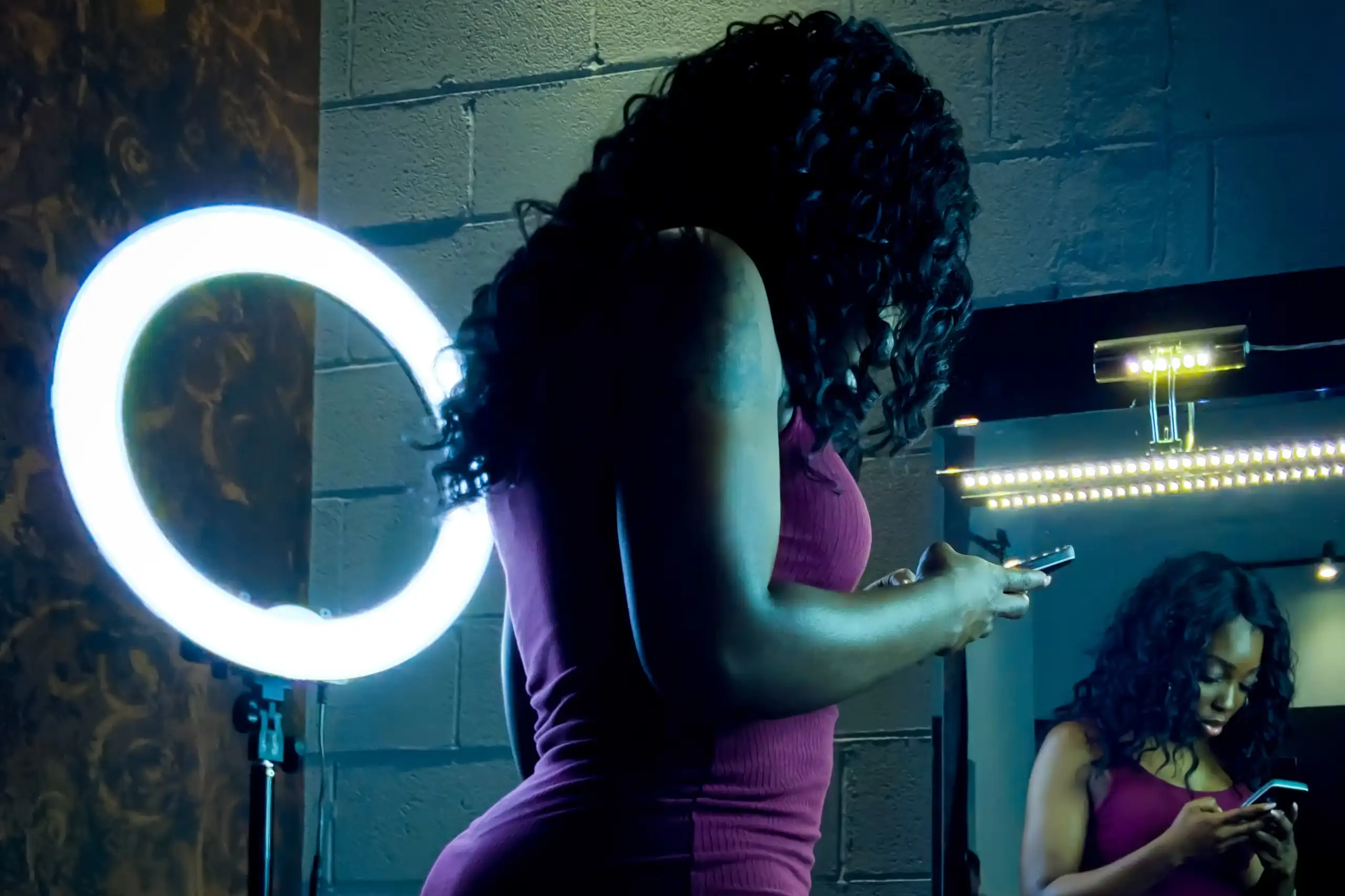 A young African woman in a tight-fitting maroon dress is reflected in a mirror as she looks at her phone, a ring light in the background.
