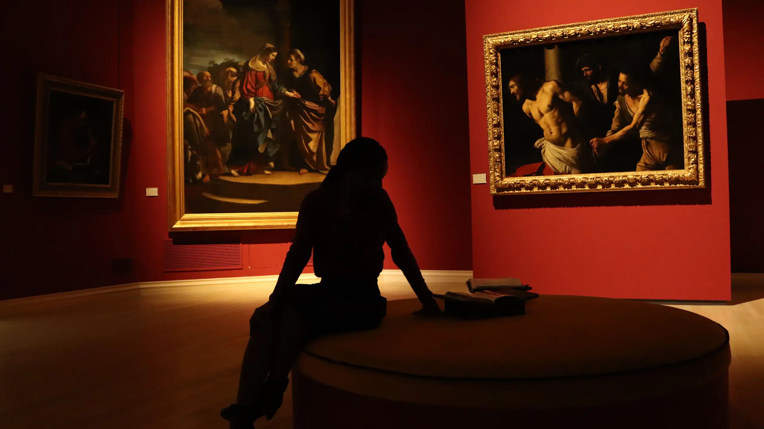 Silhouette of a woman seated in a gallery with a room decorated with red walls.