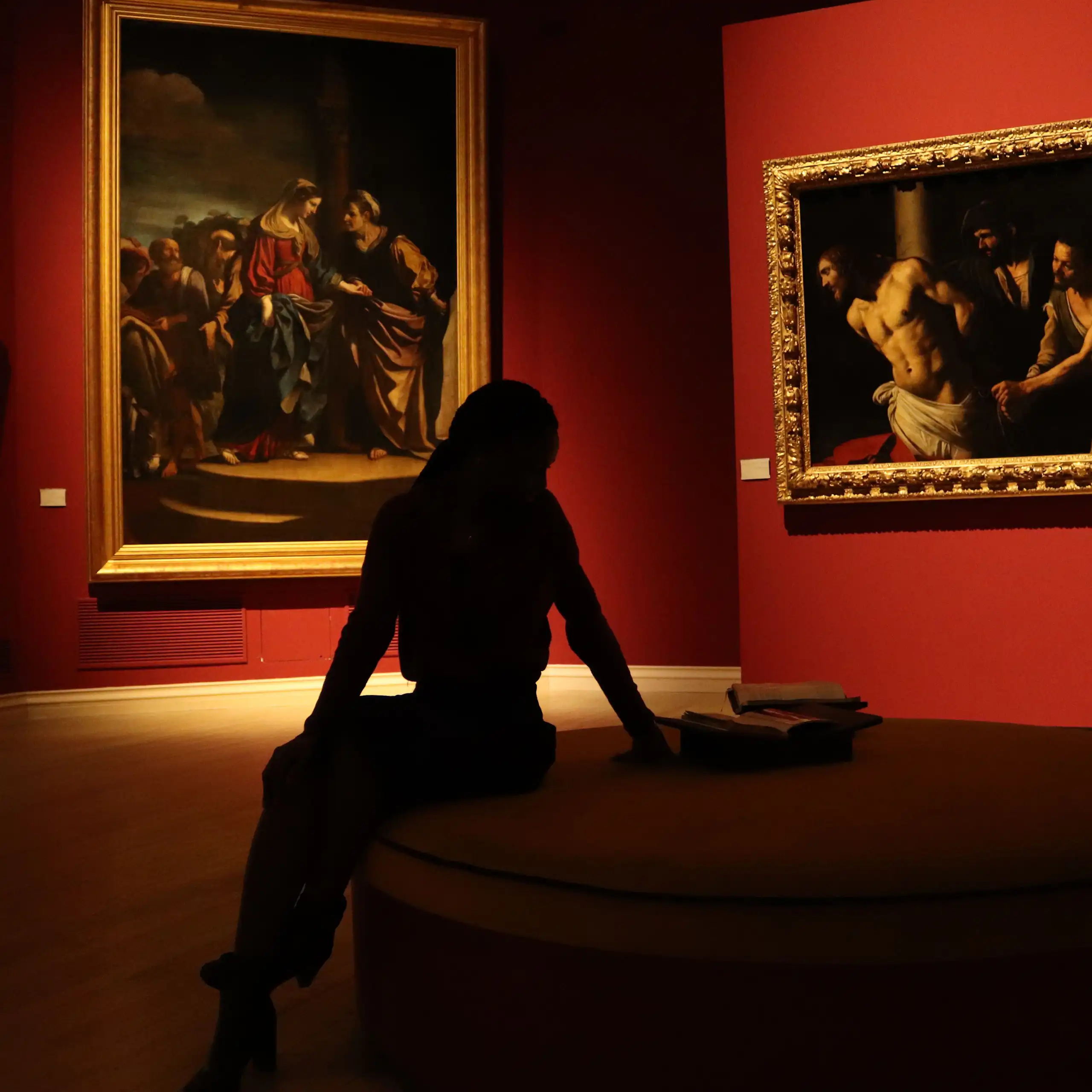 Silhouette of a woman seated in a gallery with a room decorated with red walls.