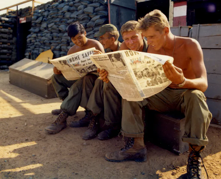 A group of young soldiers sit on crates while reading two newspapers.