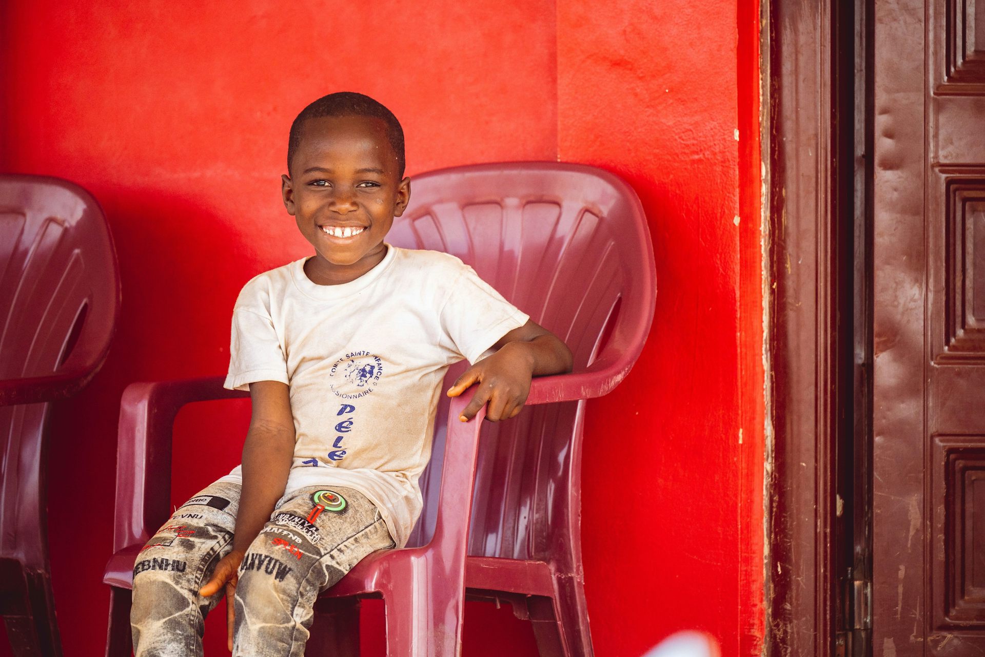 Un jeune enfant est assis sur une chaise, près d’une porte. En arrière-plan, la peinture rouge du mur est légèrement écaillée
