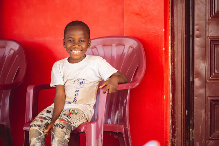 Un jeune enfant est assis sur une chaise, près d’une porte. En arrière-plan, la peinture rouge du mur est légèrement écaillée