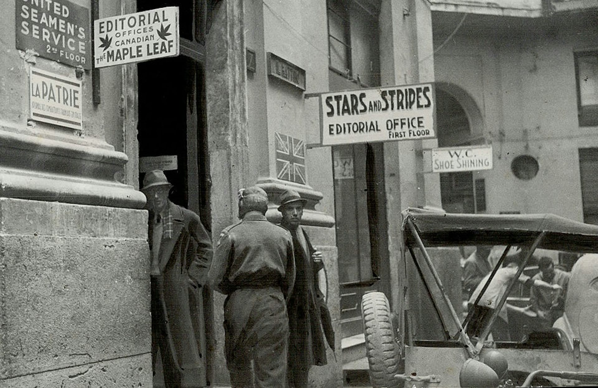 Black-and-white photo of a few men standing near a doorway, in front of signage for the Stars and Stripes editorial office.