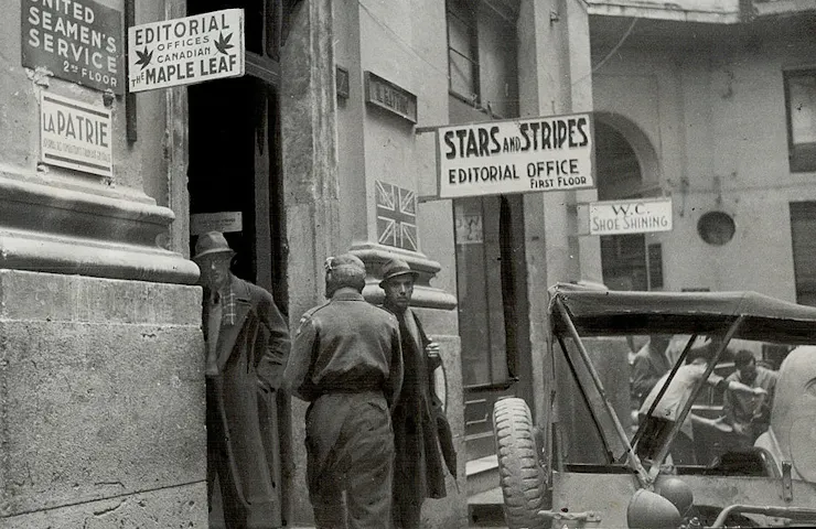 Black-and-white photo of a few men standing near a doorway, in front of signage for the Stars and Stripes editorial office.